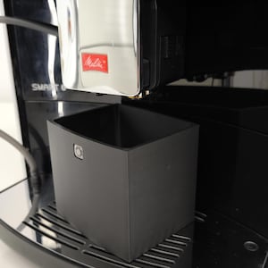 Könnte beinhalten: Eine schwarze Kaffeemaschine mit silbernen Verzierungen und einer schwarzen Tropfschale. Die Kaffeemaschine hat einen silbernen Knopf mit dem Wort "Melitta" darauf. Die Kaffeemaschine ist mit "BARISTA T1" und "SMART" beschriftet.