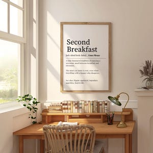 May include: A black and white printable wall art print with the definition of "Second Breakfast" in a modern minimalist style. The print is framed and hanging on a white wall above a wooden desk with books and a lamp.