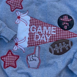 May include: Grey sweatshirt decorated with red and white gingham star and pennant appliqués. The pennant reads "GAME DAY". Other appliqués include a football, a foam finger, and a cowboy hat.