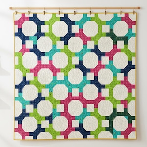 May include: A colorful quilt with a geometric pattern. The design features octagons and squares in shades of pink, green, blue, and white. The quilt is hung on a wooden rod with small clips.