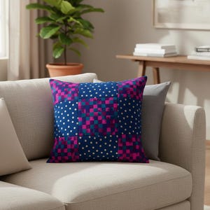 Puede incluir: Cojín cuadrado con diseño de patchwork. El cojín presenta un fondo azul marino con lunares blancos y secciones de cuadrados magenta y turquesa. El cojín está colocado sobre un sofá beige claro.