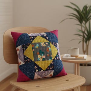 Puede incluir: Un cojín cuadrado acolchado con un diseño de patchwork. El centro presenta un rombo amarillo con detalles florales, rodeado de telas azules, moradas y estampadas. El cojín tiene un borde magenta y descansa sobre una silla de madera.