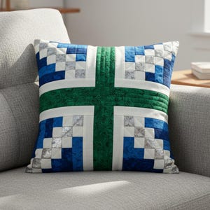 May include: Square decorative pillow with a patchwork design. The pillow features a green cross shape on a white background, with blue and silver squares on the sides. The pillow is on a gray couch.