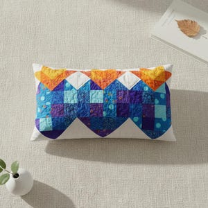 May include: Rectangular cushion with a white base and a patchwork design. The patchwork features blue, purple, and orange geometric shapes. The cushion is placed on a neutral-coloured surface.