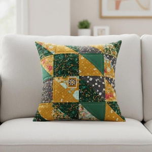 May include: Square throw pillow with a patchwork design. The pillow features a mix of fabrics in shades of green, gold, gray, and floral patterns. The pillow is placed on a white sofa.