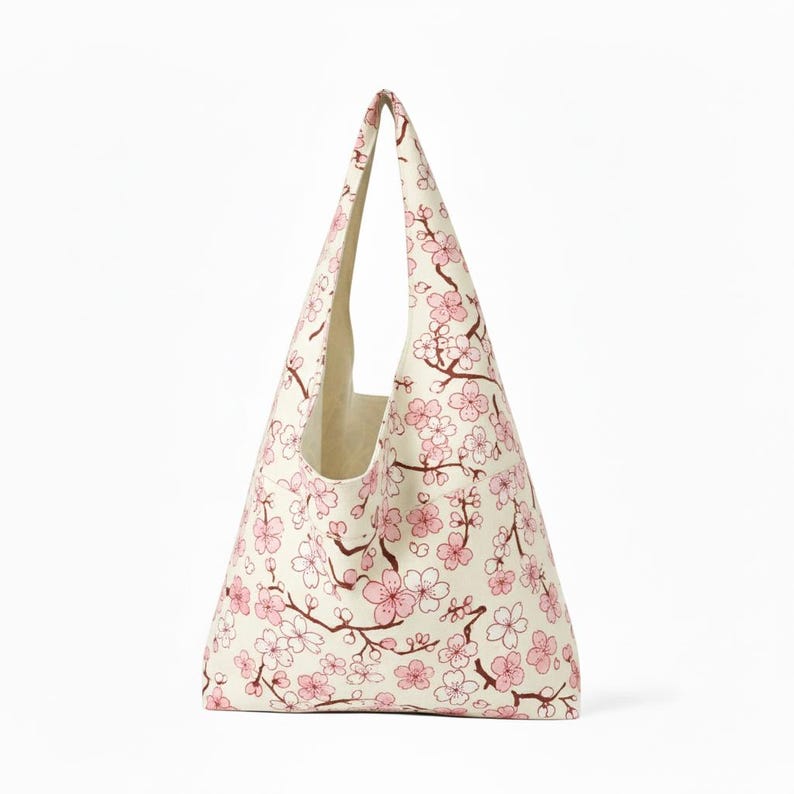 Peut inclure: Un sac fourre-tout cr&egrave;me avec un motif de fleurs de cerisier roses. Le sac a une forme triangulaire avec une seule poign&eacute;e et est fait de tissu. Le motif de fleurs de cerisier pr&eacute;sente des fleurs roses et des branches marron.