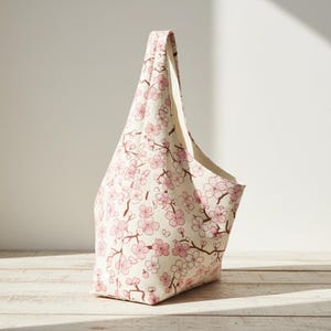 Peut inclure: Un sac fourre-tout cr&egrave;me avec un motif de fleurs de cerisier roses. Le sac a une seule poign&eacute;e et une forme triangulaire. Le motif floral pr&eacute;sente des fleurs roses et des branches marron.