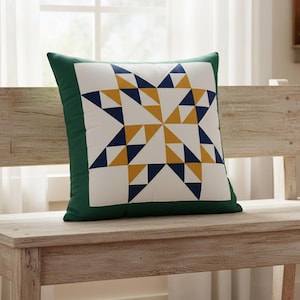 Op de afbeelding: Vierkant decoratief kussen met een ster quiltpatroon. Het kussen heeft een wit midden met een sterontwerp in marineblauwe en gouden driehoeken. De rand is effen donkergroen.