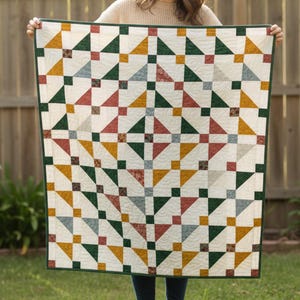 Könnte beinhalten: Eine handgefertigte Steppdecke mit einem geometrischen Muster. Die Steppdecke zeigt Dreiecke und Quadrate in Grün-, Gold-, Rot- und Grautönen auf cremefarbenem Hintergrund. Die Steppdecke ist mit einem grünen Rand versehen.