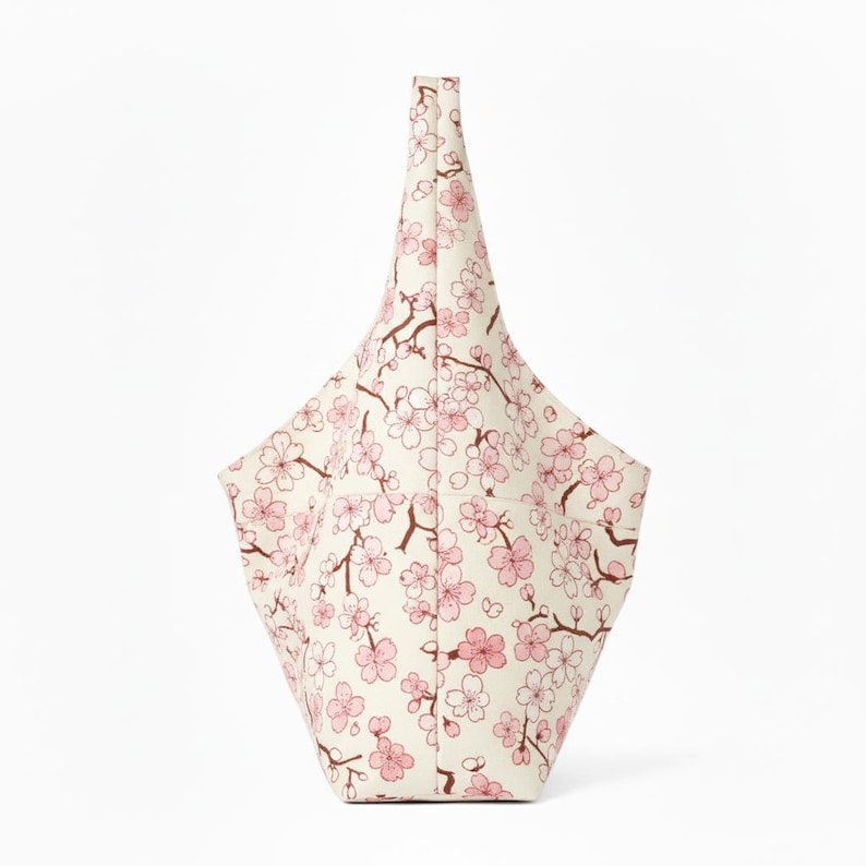 Peut inclure: Un sac fourre-tout cr&egrave;me avec un motif de fleurs de cerisier roses. Le sac a une forme angulaire unique avec une poign&eacute;e haute et pointue. Le motif floral pr&eacute;sente des fleurs roses et des branches marron.