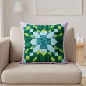 May include: A decorative throw pillow with a geometric design. The pillow features a central teal pattern with a dotted design, surrounded by green, light blue, and yellow geometric shapes. The pillow has a lavender border and is placed on a beige sofa.
