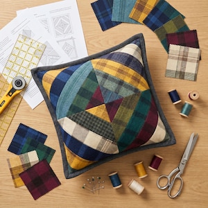 Puede incluir: Un cojín de patchwork con un diseño geométrico en varias telas de cuadros, incluyendo azul, verde, amarillo y burdeos. Suministros de costura como tijeras, carretes de hilo y muestras de tela están dispuestos alrededor del cojín.