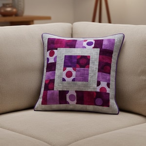 Op de afbeelding: Vierkant decoratief kussen met een patchwork ontwerp in tinten paars, magenta en grijs. Het kussen heeft een centraal vierkant met een stippenpatroon en een grijze rand, met een paarse rand.