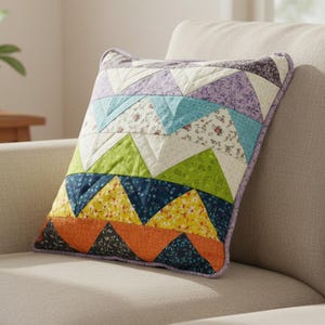 May include: A colorful, patchwork pillow with a geometric design. The pillow features triangles in various colors, including orange, yellow, green, blue, and purple. The pillow is approximately 16 inches square and has a lavender border.