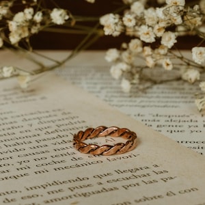 Op de afbeelding: Een koperkleurige gevlochten ring ligt op een vintage pagina, met delicate witte bloemen op de achtergrond. Het ontwerp van de ring heeft verweven strengen, waardoor een textuur ontstaat. De ring is een sieraad.