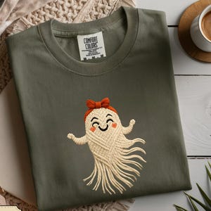 May include: A moss green t-shirt featuring a smiling ghost design with a red bow and macrame-style body. The shirt has a crew neck and short sleeves. The text "COMFORT COLORS" is visible on the tag. The image also includes the text "High-quality direct-to-garment (DTG) print!"