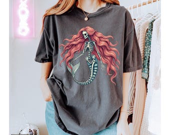 May include: A charcoal gray short-sleeve t-shirt featuring a graphic print of a skeleton mermaid with long, flowing pink hair. The skeleton has a rib cage, arms, and a fish tail. The shirt has a crew neckline and a relaxed, loose-fitting silhouette.