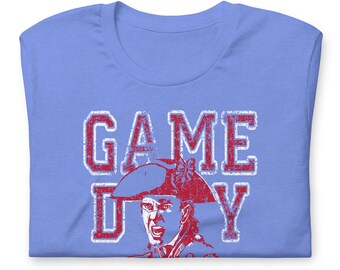 May include: A light blue t-shirt with the words "GAME DAY" in distressed red lettering. Below the text is a red illustration of a person wearing a tricorn hat, with an open mouth.