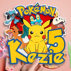 May include: A colorful Pok&eacute;mon-themed birthday cake topper featuring Pikachu, other characters, and the name "Kezie" with the number "5". The design includes a red background with the Pok&eacute;mon logo and various cartoon characters.