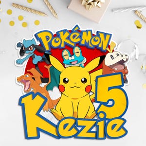 May include: A colorful birthday cake topper featuring the word "Pok&eacute;mon" in yellow and blue, with a red background. The number "5" and the name "Kezie" are also displayed. Several Pok&eacute;mon characters are included in the design.