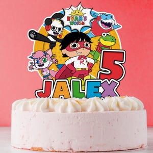 Puede incluir: Un pastel rosa glaseado cubierto con un adorno de pastel colorido con personajes de dibujos animados y el texto "RYAN'S WORLD". El adorno incluye el número "5" y el nombre "JALEX". El pastel está sobre una superficie blanca sobre un fondo rosa.