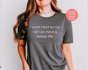 May include: A dark gray t-shirt with the text "I work hard so my cat can have a better life" printed in white. The shirt is a unisex style.