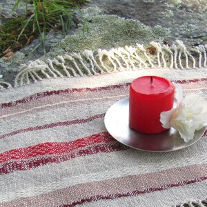 Handcrafted Small Positive Energy Red Altar Cloth Table Mat, Handmade Artisan Woven Spiritual Meditation, Reiki Root Chakra, Fire Element Feng Shui, Fire Red Fabric Table Centerpiece by Amy C. Lund in Burgundy Red, Blush Rose and Beige Gray with striped bands of alternating color and texture patterns. Styled on a rock surface with a red candle is sitting on a silver plate and white flower.