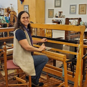 Amy C. Lund Handweaver and fiber artist, sitting at a loom in the weaving studio and gallery where she teaches and creates artisan handspun yarns from sheep's wool, alpaca, cotton, linen and other fibers; hand knit and hand woven accessories including scarves, handbags and clothing; home decor table linens, blankets, rugs, tapestries; mixed media fiber art collage assemblages; and repairs textiles. She has a M.S. degree in Textiles & Related Art from the University of Rhode Island in 1993.