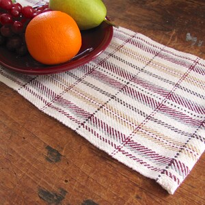 Puede incluir: Un camino de mesa blanco con un patrón de cuadros en tonos burdeos, gris y beige. El camino de mesa está sobre una mesa de madera con un plato de fruta encima.