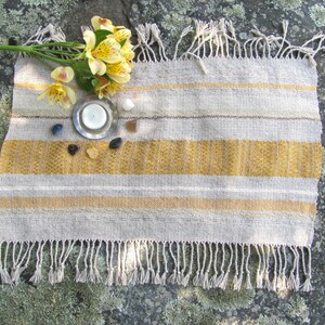 Small Altar Cloth Table Mat Centerpiece, Artisan Handmade Hand Woven Sun Yellow, Brown, Ivory White and Beige Gray Handcrafted by Amy C. Lund with striped bands of color and textural patterns and hand knotted fringe.  Styled with a small, white candle on a metal plate circled by small, colorful stones, and yellow freesia on a stone surface.