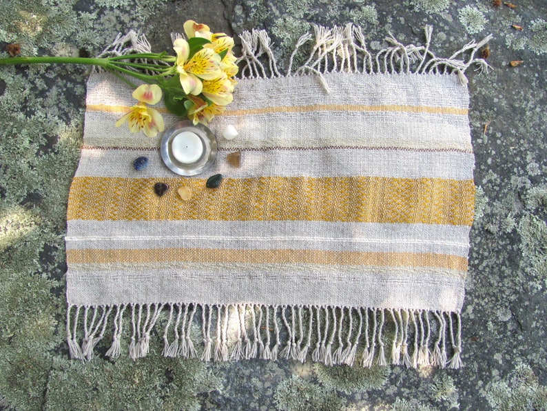 Small Altar Cloth Table Mat Centerpiece, Artisan Handmade Hand Woven Sun Yellow, Brown, Ivory White and Beige Gray Handcrafted by Amy C. Lund with striped bands of color and textural patterns and hand knotted fringe.  Styled with a small, white candle on a metal plate circled by small, colorful stones, and yellow freesia on a stone surface.