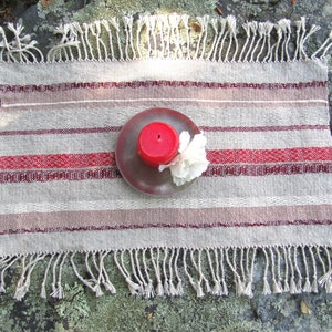 Handcrafted Small Positive Energy Red Altar Cloth Table Mat, Handmade Artisan Woven Spiritual Meditation, Reiki Root Chakra, Fire Element Feng Shui, Fire Red Fabric Table Centerpiece by Amy C. Lund in Burgundy Red, Blush Rose and Beige Gray with striped bands of alternating color and texture patterns. Styled on a rock surface with a red candle is sitting on a silver plate and white flower.