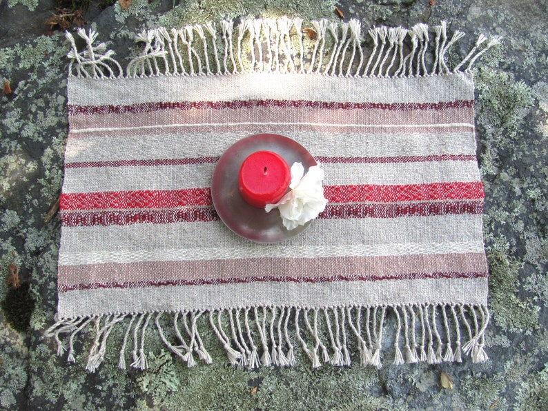 Handcrafted Small Positive Energy Red Altar Cloth Table Mat, Handmade Artisan Woven Spiritual Meditation, Reiki Root Chakra, Fire Element Feng Shui, Fire Red Fabric Table Centerpiece by Amy C. Lund in Burgundy Red, Blush Rose and Beige Gray with striped bands of alternating color and texture patterns. Styled on a rock surface with a red candle is sitting on a silver plate and white flower.