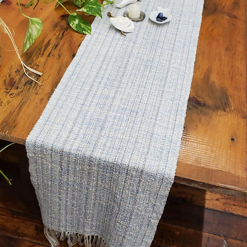 Handmade Light Blue Chalcedony Artisan Hand Woven Scarf Shawl or Table Runner for Positive Natural Energy, Spiritual Serenity, Harmony, Grounding Meditation Prayer is a Gender Neutral Unisex Men's Women's Long Fall, Winter, Spring, Urban Casual Rustic Fashion Accessory Wrap or Home Decor Accent Handcrafted by Amy C. Lund in Cotton Blend. Styled on a wood table with a plant, white fish shaped dish with blue stones, a small white candle on a metal dish, a seashell.
