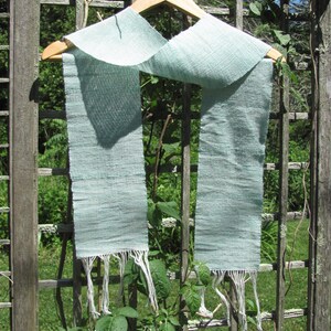 May include: A light blue woven scarf with white fringe hanging on a wooden hanger against a wooden lattice background.