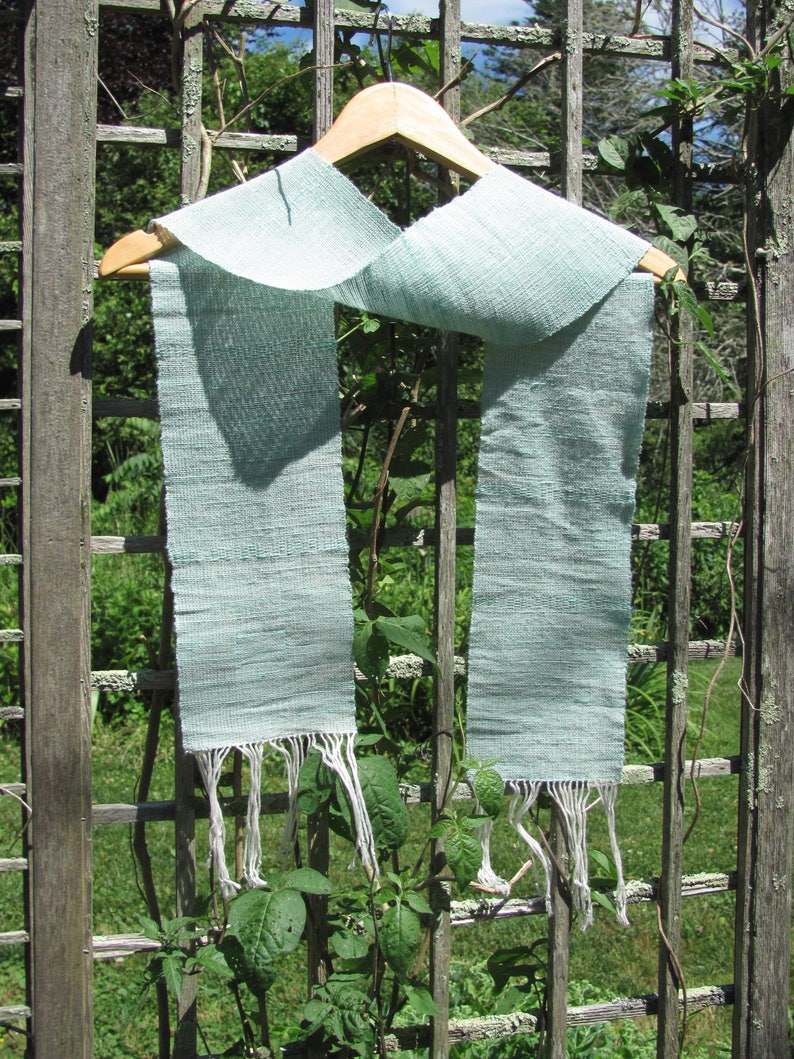 May include: A light blue woven scarf with white fringe hanging on a wooden hanger against a wooden lattice background.