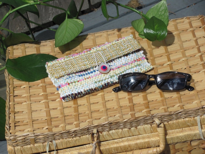 Boho Wallet Smart Phone Case, Small Hand Woven Clutch Purse, Cloth Fabric Soft Eyeglass Case Handmade Vegan Eco Friendly Zero Waste Upcycled Recycled Bag, Sustainable Slow Fashion Accessory with Sunny Yellow Accents Handcrafted by Amy C. Lund! Showing button closure. Styled with sunglasses on a basket with green leaves.