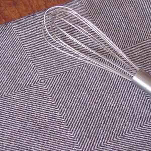 A Navy Blue and White Hand Woven Twill Block Pattern kitchen towel, shown as an example for Table or Floor Loom Weaving Pattern Draft PDF, 4 H Harness Shaft Handweaving Cotton Kitchen Hand Woven Dish Tea Towel Downloadable Tutorial, an artful Kitchenware, Cookware Accessory for Gourmet Chef, Home Baking, Cooking, Instructions Designed by Master Weaver Amy C. Lund. Pictured with a silver metal whisk with a handle.