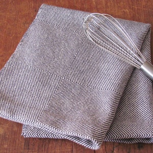 A folded navy blue and white Hand Woven Twill Block Pattern kitchen towel, shown as an example for Table or Floor Loom Weaving Pattern Draft PDF, 4 H Harness Shaft Handweaving Cotton Kitchen Hand Woven Dish Tea Towel Downloadable Tutorial, an artful Kitchenware, Cookware Accessory for Gourmet Chef, Home Baking, Cooking, Instructions Designed by Master Weaver Amy C. Lund. A silver whisk is lying on top of the towels.