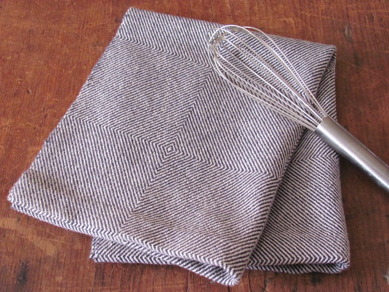A folded navy blue and white Hand Woven Twill Block Pattern kitchen towel, shown as an example for Table or Floor Loom Weaving Pattern Draft PDF, 4 H Harness Shaft Handweaving Cotton Kitchen Hand Woven Dish Tea Towel Downloadable Tutorial, an artful Kitchenware, Cookware Accessory for Gourmet Chef, Home Baking, Cooking, Instructions Designed by Master Weaver Amy C. Lund. A silver whisk is lying on top of the towels.