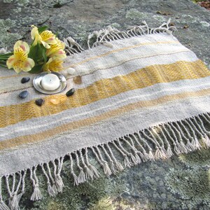 Small Altar Cloth Table Mat Centerpiece, Artisan Handmade Hand Woven Sun Yellow, Brown, Ivory White and Beige Gray Handcrafted by Amy C. Lund with striped bands of color and textural patterns and hand knotted fringe.  Styled with a small, white candle on a metal plate circled by small, colorful stones, and yellow freesia on a stone surface.