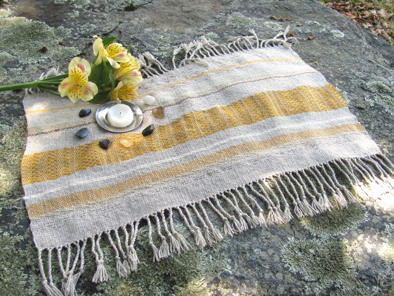 Small Altar Cloth Table Mat Centerpiece, Artisan Handmade Hand Woven Sun Yellow, Brown, Ivory White and Beige Gray Handcrafted by Amy C. Lund with striped bands of color and textural patterns and hand knotted fringe.  Styled with a small, white candle on a metal plate circled by small, colorful stones, and yellow freesia on a stone surface.