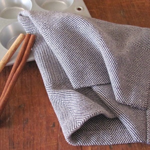 A Navy Blue and White Hand Woven Twill Block Pattern kitchen towel, shown as an example for Table or Floor Loom Weaving Pattern Draft PDF, 4 H Harness Shaft Handweaving Cotton Kitchen Hand Woven Dish Tea Towel Downloadable Tutorial, an artful Kitchenware, Cookware Accessory for Gourmet Chef, Home Baking, Cooking, Instructions Designed by Master Weaver Amy C. Lund.  Pictured with two cinnamon sticks next to a silver muffin tin.