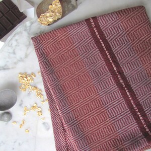Handcrafted Kitchen Dish Towel, Gourmet Baking, Home Cooking gift for Chef, Baker, Cook, Foodie. An Eco Sustainable, Reusable, Artisan Handmade Cookware Kitchenware Accessory Hand Woven Cotton Towel by Amy C. Lund in Deep Burgundy Red, Beige, White & Fudge Brown Stripe Cloth for Rustic French Country or Modern Farmhouse Decor with alternating Chevron Diamond Zig Zag Herringbone. Styled folded on a white marble surface with a bar of chocolate, metal measuring spoons, and oats.