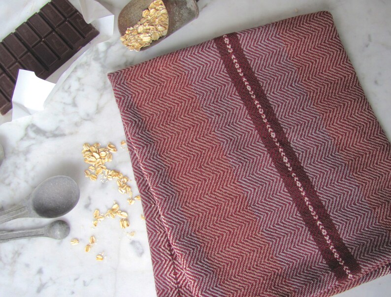 Handcrafted Kitchen Dish Towel, Gourmet Baking, Home Cooking gift for Chef, Baker, Cook, Foodie. An Eco Sustainable, Reusable, Artisan Handmade Cookware Kitchenware Accessory Hand Woven Cotton Towel by Amy C. Lund in Deep Burgundy Red, Beige, White & Fudge Brown Stripe Cloth for Rustic French Country or Modern Farmhouse Decor with alternating Chevron Diamond Zig Zag Herringbone. Styled folded on a white marble surface with a bar of chocolate, metal measuring spoons, and oats.
