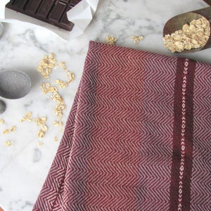 Handcrafted Kitchen Dish Towel, Gourmet Baking, Home Cooking gift for Chef, Baker, Cook, Foodie. An Eco Sustainable, Reusable, Artisan Handmade Cookware Kitchenware Accessory Hand Woven Cotton Towel by Amy C. Lund in Deep Burgundy Red, Beige, White & Fudge Brown Stripe Cloth for Rustic French Country or Modern Farmhouse Decor with alternating Chevron Diamond Zig Zag Herringbone. Styled on a white marble surface with a bar of chocolate, metal measuring spoons, and oats.