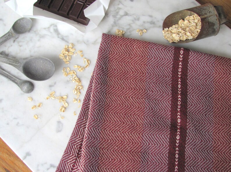 Handcrafted Kitchen Dish Towel, Gourmet Baking, Home Cooking gift for Chef, Baker, Cook, Foodie. An Eco Sustainable, Reusable, Artisan Handmade Cookware Kitchenware Accessory Hand Woven Cotton Towel by Amy C. Lund in Deep Burgundy Red, Beige, White & Fudge Brown Stripe Cloth for Rustic French Country or Modern Farmhouse Decor with alternating Chevron Diamond Zig Zag Herringbone. Styled on a white marble surface with a bar of chocolate, metal measuring spoons, and oats.