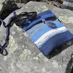 Small Crossbody Bag Wallet or Phone Purse is Handmade, Artisan Hand Woven in Indigo Navy Blue with Zero Waste, Eco Friendly Upcycled Cloth, Vegan Recycled Fabric. This Sustainable Slow Fashion Accessory Handbag, Boho Cash Change Pouch or Money Shoulder Pouch is Handcrafted by Amy C. Lund. Styled casually with black sunglasses on a gray rock.