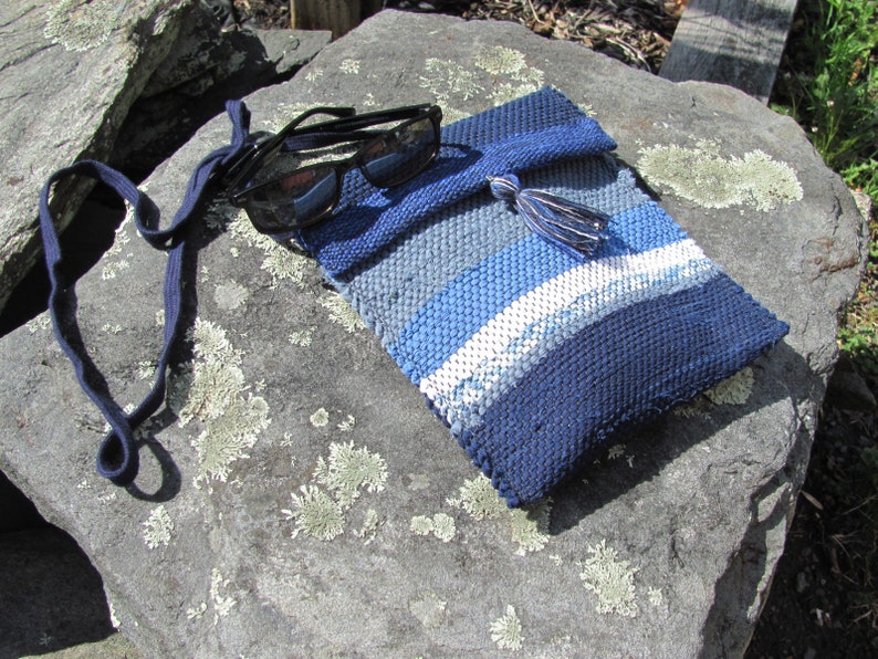 Small Crossbody Bag Wallet or Phone Purse is Handmade, Artisan Hand Woven in Indigo Navy Blue with Zero Waste, Eco Friendly Upcycled Cloth, Vegan Recycled Fabric. This Sustainable Slow Fashion Accessory Handbag, Boho Cash Change Pouch or Money Shoulder Pouch is Handcrafted by Amy C. Lund. Styled casually with black sunglasses on a gray rock.