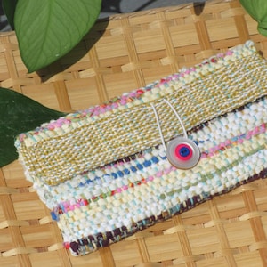 Boho Wallet Smart Phone Case, Small Hand Woven Clutch Purse, Cloth Fabric Soft Eyeglass Case Handmade Vegan Eco Friendly Zero Waste Upcycled Recycled Bag, Sustainable Slow Fashion Accessory with Sunny Yellow Accents Handcrafted by Amy C. Lund! Showing button closure. Styled on a basket with green leaves.